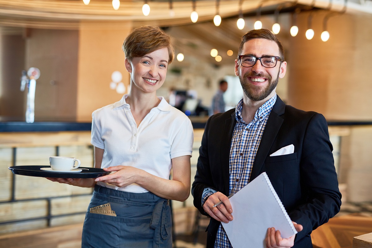 Group portrait of cheerful bearded restaurant manager, and pretty waitress posing for photography while standing against bar counter illustrating the success restaurant operators can have when hiring restaurant employees through WOTC.